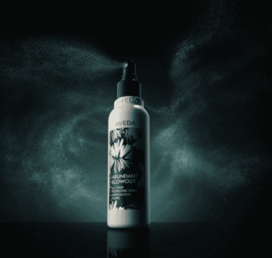 A bottle of Aveda Abundant Blowout hair styling spray stands upright, mist spraying from its nozzle against a dark, smoky background. | AMORE Salon, St. Petersburg, FL