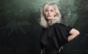 A woman with platinum blonde hair wearing a black outfit stands in front of a dark green, textured background, looking directly at the camera. | AMORE Salon, St. Petersburg, FL