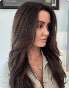 Woman with long, layered brown hair wearing a light-colored shirt and a green pendant necklace, looking to the side in a well-lit indoor setting. | AMORE Salon, St. Petersburg, FL