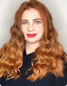 A woman with long, wavy, auburn hair and fair skin is wearing a black top and smiling slightly against a plain light background. | AMORE Salon, St. Petersburg, FL