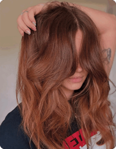 A person with long, wavy, reddish-brown hair partially covering their face holds one hand on top of their head. | AMORE Salon, St. Petersburg, FL