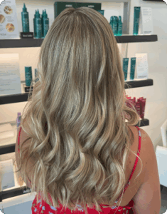 Person with long, wavy blonde hair wearing a red floral top, standing in front of shelves with hair products at a salon. | AMORE Salon, St. Petersburg, FL