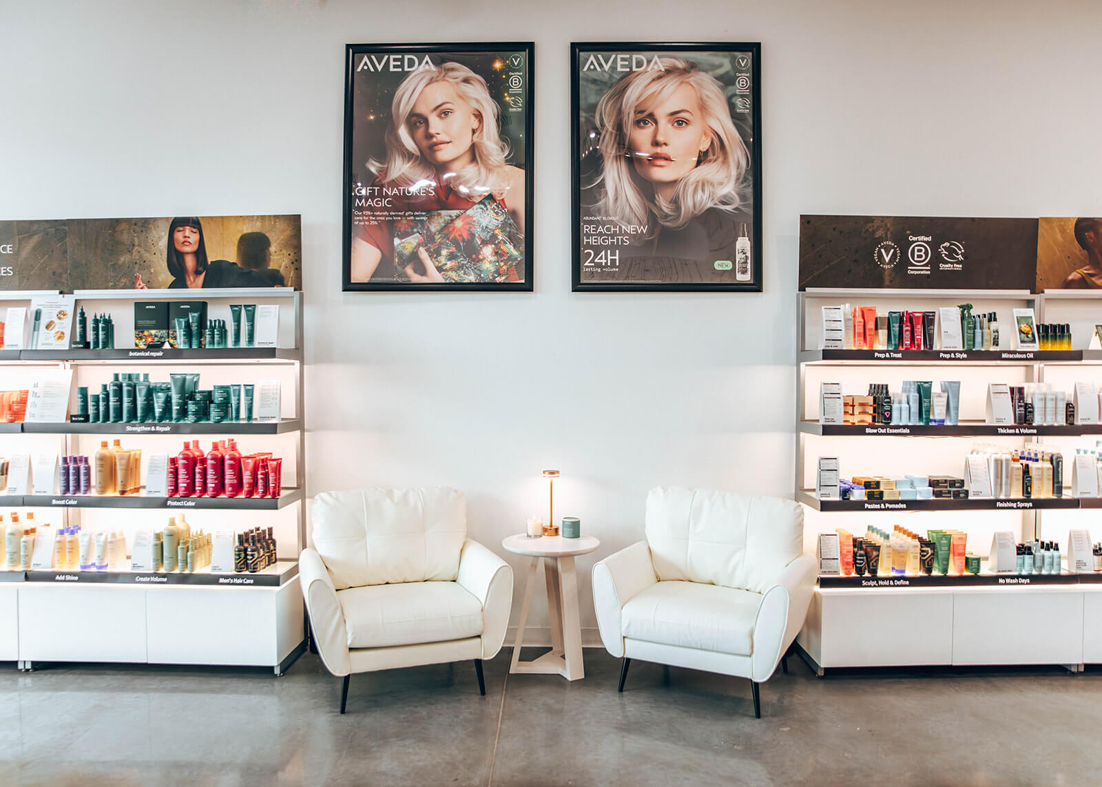 Two white chairs and a small table are positioned between two shelves displaying hair and beauty products, with two large Aveda posters on the wall above. | AMORE Salon, St. Petersburg, FL