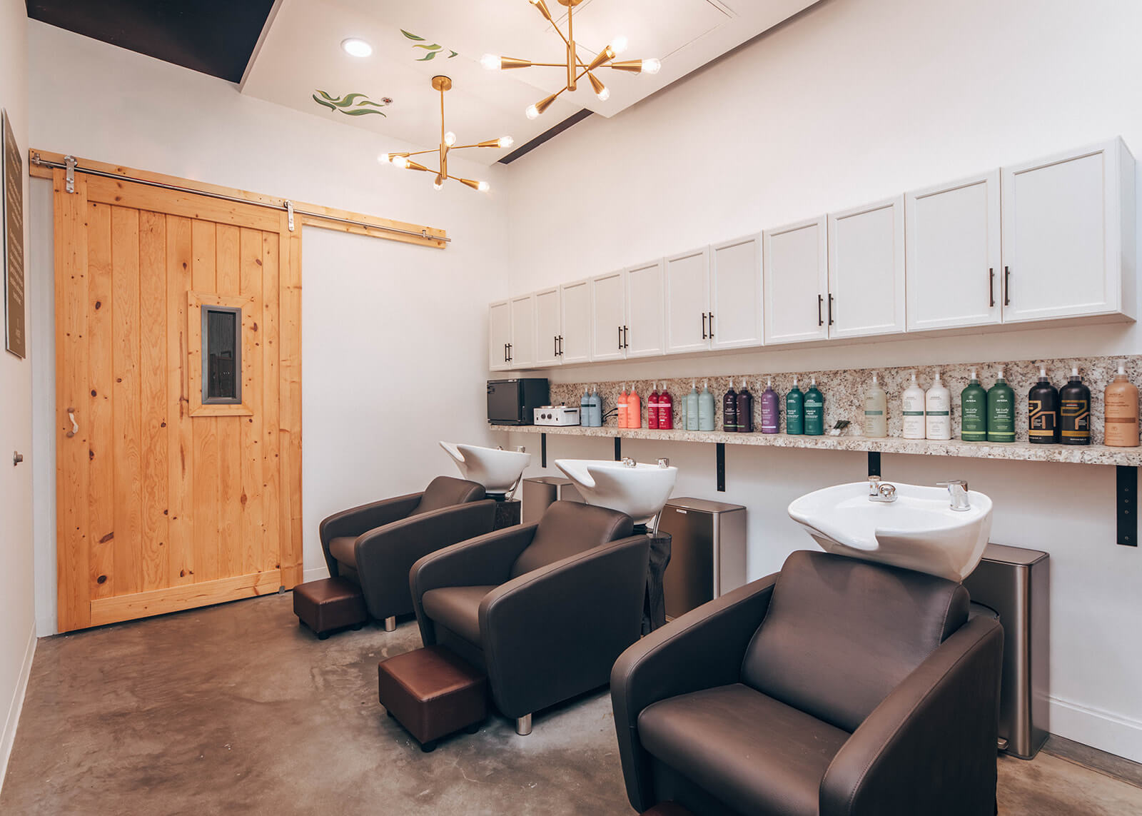 Three black salon shampoo chairs with white sinks face a wall with white cabinets and colorful shampoo bottles; wooden sliding door and modern lighting are visible. | AMORE Salon, St. Petersburg, FL