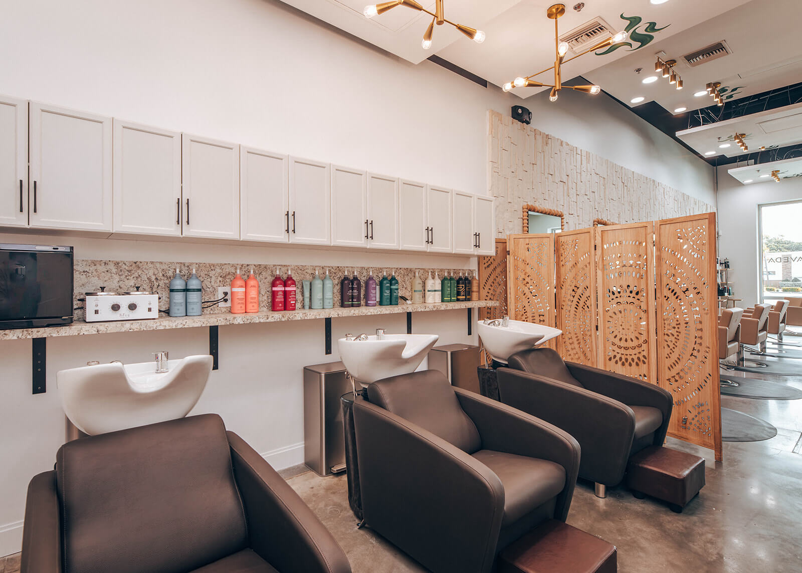 Modern hair salon with three brown shampoo chairs, white sinks, shelves with hair products, and decorative wooden divider panels. | AMORE Salon, St. Petersburg, FL