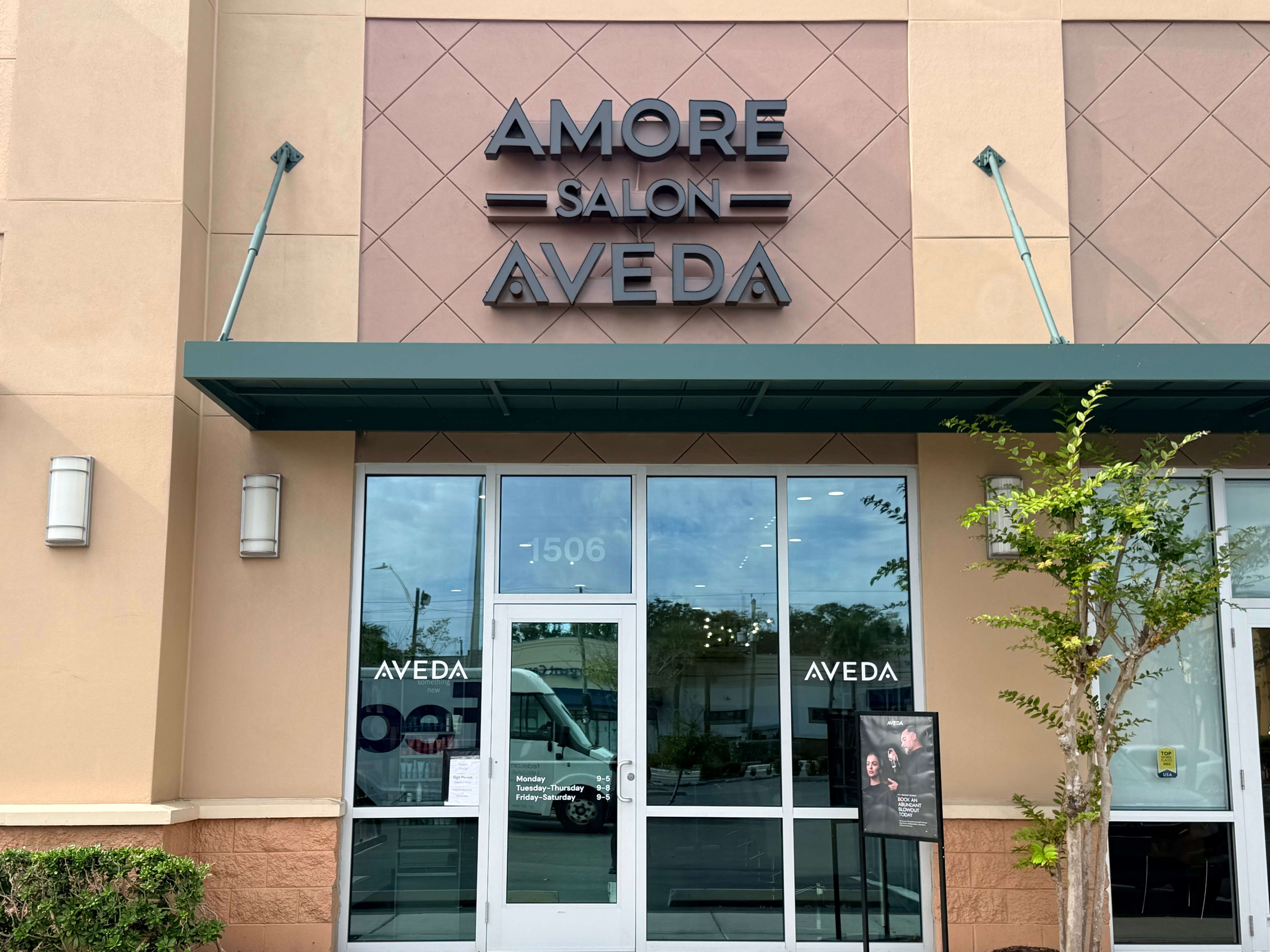 Storefront of Amore Salon Aveda with glass doors and windows, a sign above the entrance, and a small tree to the right of the entrance. | AMORE Salon, St. Petersburg, FL