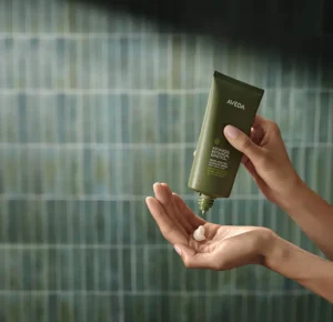 A person dispenses a botanical skincare cream from a green Aveda tube onto their palm in front of a tiled wall. | AMORE Salon, St. Petersburg, FL