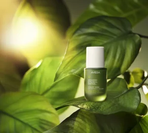 A small green bottle of Aveda Advanced Botanical Kinetics Vitality Serum, featuring vegan peptides, is placed on large green leaves with sunlight in the background. | AMORE Salon, St. Petersburg, FL