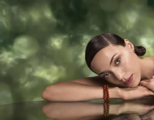 A woman with sleek dark hair rests her head and arms on a reflective surface, wearing a red beaded bracelet, embodying the natural glow inspired by botanical skincare, with a blurred green background. | AMORE Salon, St. Petersburg, FL