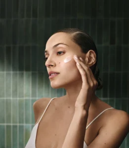 A woman applies cream to her cheek with her fingers, standing in front of green tiled wall, wearing a light-colored camisole. | AMORE Salon, St. Petersburg, FL