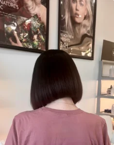 Person with straight, dark bob haircut seen from behind, wearing a mauve shirt, inside a salon with beauty product posters and shelves visible on the wall. | AMORE Salon, St. Petersburg, FL