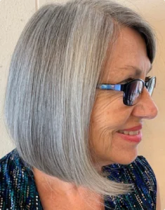 Older woman with straight, chin-length gray hair and glasses, smiling and looking to the side. She is wearing a blue patterned top. | AMORE Salon, St. Petersburg, FL