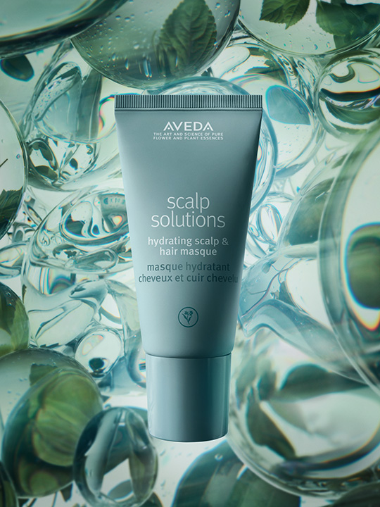 A grey tube of Aveda Scalp Solutions hydrating scalp and hair masque stands upright against a background of water droplets and green leaves, offering a boost for scalp health recommended by any expert hair stylist. | AMORE Salon, St. Petersburg, FL