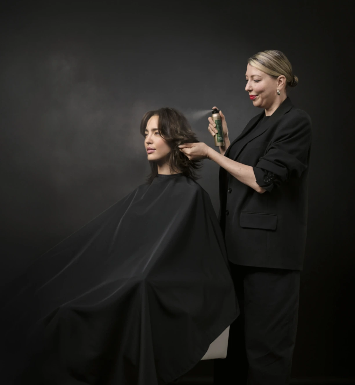 A hairstylist dressed in black sprays product on a seated client's hair against a dark background. | AMORE Salon, St. Petersburg, FL