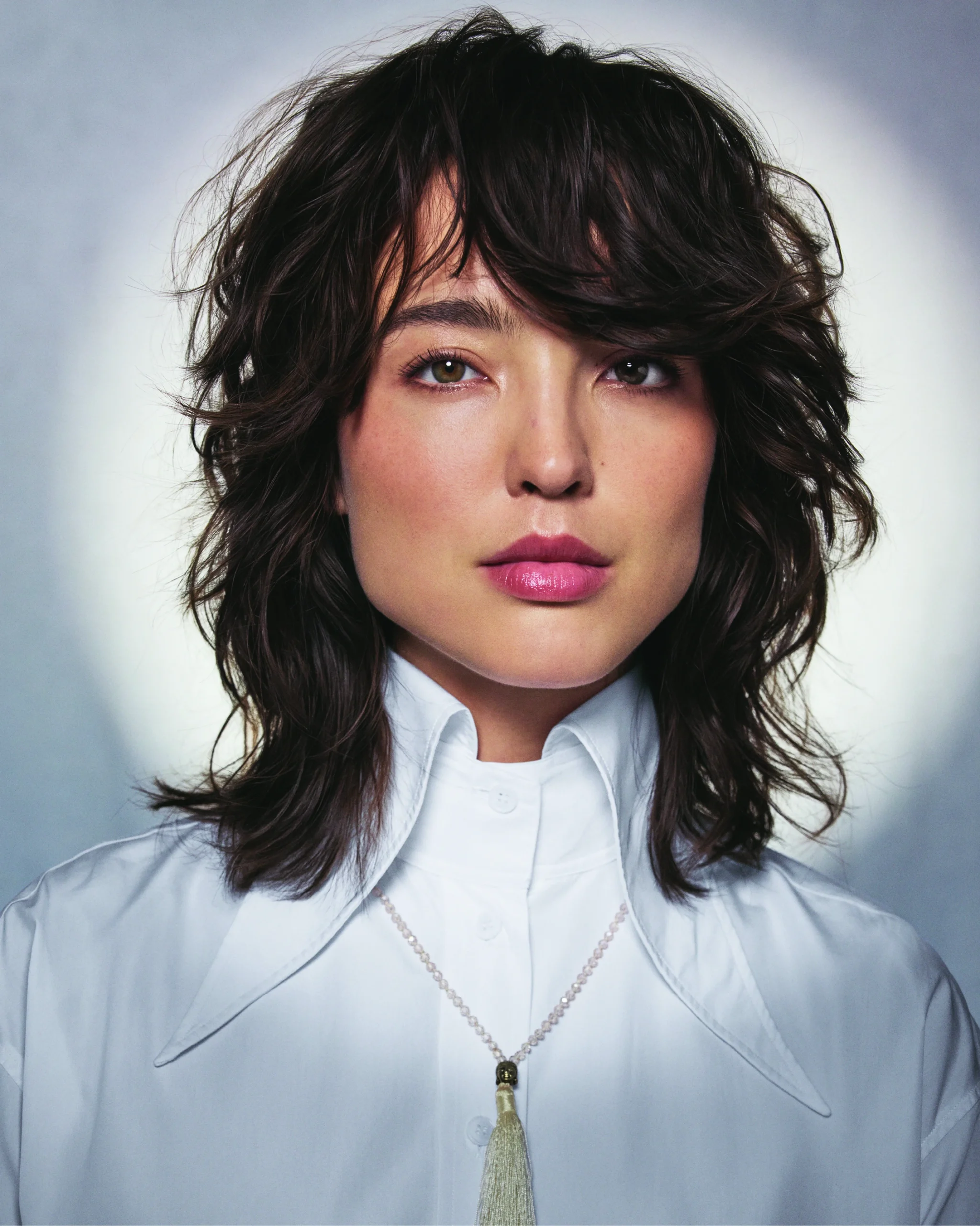 A person with wavy brown hair wearing a white collared shirt and a necklace with a tassel pendant, posed against a soft gradient background. | AMORE Salon, St. Petersburg, FL