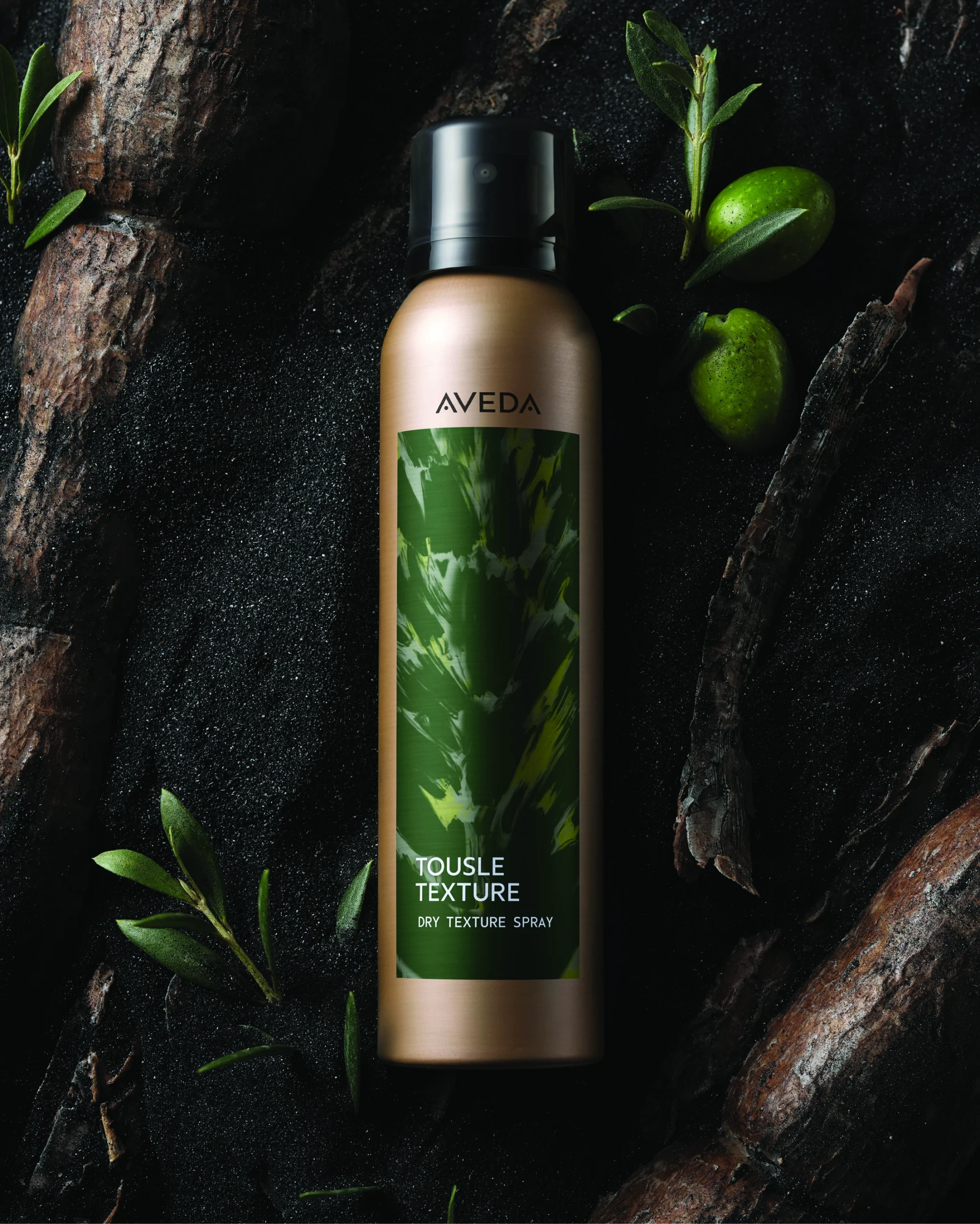 Aveda Tousle Texture dry texture spray canister on a dark surface, surrounded by green olives, leaves, and bark. | AMORE Salon, St. Petersburg, FL